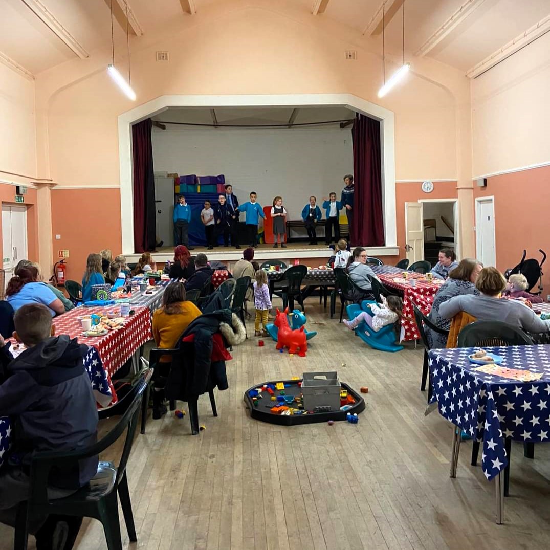 A photo of Free Tea Friday, a horse shoe of tables in front of the stage with children and Revd-Rachel saying the prayers.